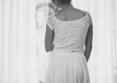 Black & White photo of the back of bride in a wedding dress looking out the window, while holding back the soft lace curtains