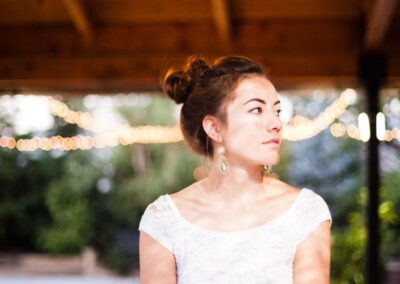 A brunette bride with hair up in white lace wedding gown under the wooden Pavilion with backdrop lights looking to the side.