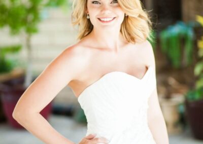 Blonde Bride wearing a lace headband standing in Courtyard in a white strapless wedding dress, smiling with right arm on hip