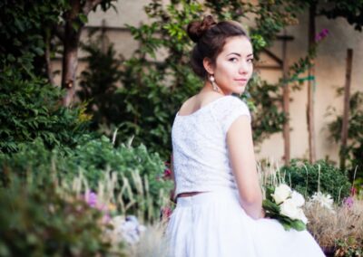 Bride with brown hair up in a bun in her white wedding gown sitting in the green lush garden, looking back at the camera.